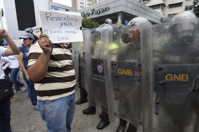 A demonstrator holds a sign reading "Recall now! The people rule 2016" in front of a line of policemen during a protest against new emergency powers decreed this week by President Nicolas Maduro in Caracas on May 18, 2016. Public outrage was expected to spill onto the streets of Venezuela Wednesday, with planned nationwide protests marking a new low point in Maduro's unpopular rule. / AFP PHOTO / JUAN BARRETO